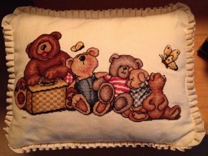 The picture shows a pillow with a cross stitched scene of five teddy bears with a picnic basket and two butterflies.