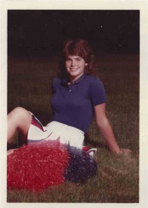 Picture of a girl sitting on the ground in a cheerleading uniform with orange and blue pompoms.