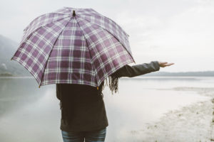 Woman standing, facing a beach, with a checkered umbrella unfolded over her shoulder and her hand out to the side checking for rain.